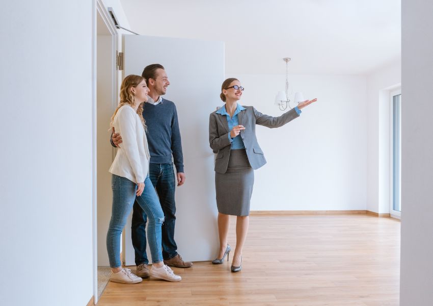 A property manager in a skirt and suit jacket shows two new tenants the inside of a clean and white rental unit.